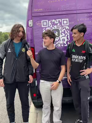 Three young men are standing in front of a purple van. The man in the middle holds a microphone, appearing to be conducting an interview. The van has a large QR code and writing on it.
