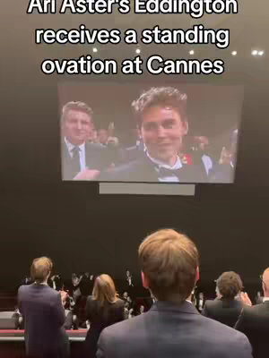 The image shows a large screen displaying a shot of two people, with a man on the right in a suit and another man in the background with a tie. In front of the screen, an audience is visible from the back, many wearing suits and clapping. At the top is black text.