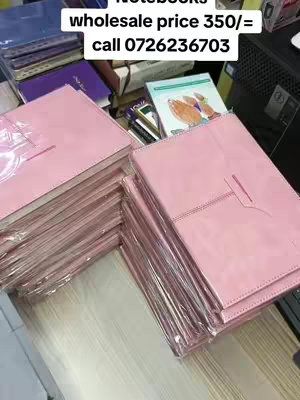 The image shows stacks of pink notebooks, some wrapped in clear plastic. The notebooks are on a wooden surface. In the background, there's a glimpse of more books and items. Text overlayed on the image provides information about wholesale prices and a phone number.