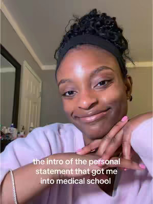 A young Black woman is centered in a selfie-style shot. She has curly hair styled with a black headband. Her skin is clear, and she wears a light pink sweater/top. She smiles slightly, resting her hands and pink polished nails beneath her chin as if deep in thought. The background is blurred, with glimpses of a mirror, a door, and some objects on a surface. Text overlayed on the image states 'the intro of the personal statement that got me into medical school'.