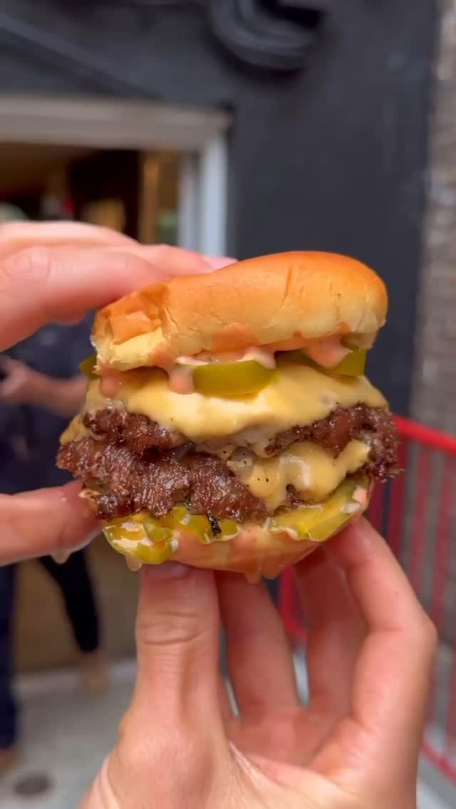 A close-up shot of a generously stacked burger held between two hands against a blurred background. The burger has a sesame seed bun, two visible beef patties, melted cheese, pickles, and a pink sauce. The lighting is natural, casting soft shadows.
