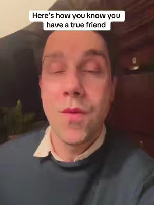 A man with fair skin, dark hair, and closed eyes is in the center of the frame. He's wearing a dark blue sweater with a white collared shirt underneath. The background is dimly lit with warm tones. Above him, a white text box reads, "Here's how you know you have a true friend."