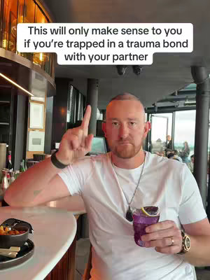 A man with a bald or shaved head, wearing a white t-shirt, is holding a purple cocktail with a lemon slice. He has a serious expression and is pointing upwards with one finger. He is seated at a bar, and the background shows other people and bar decor.