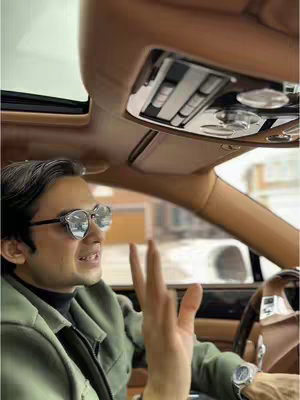 A man is seated in a car, looking towards the camera. He is wearing sunglasses and raising his hand, appearing to be talking. The car's interior is visible, showcasing the roof and dashboard.