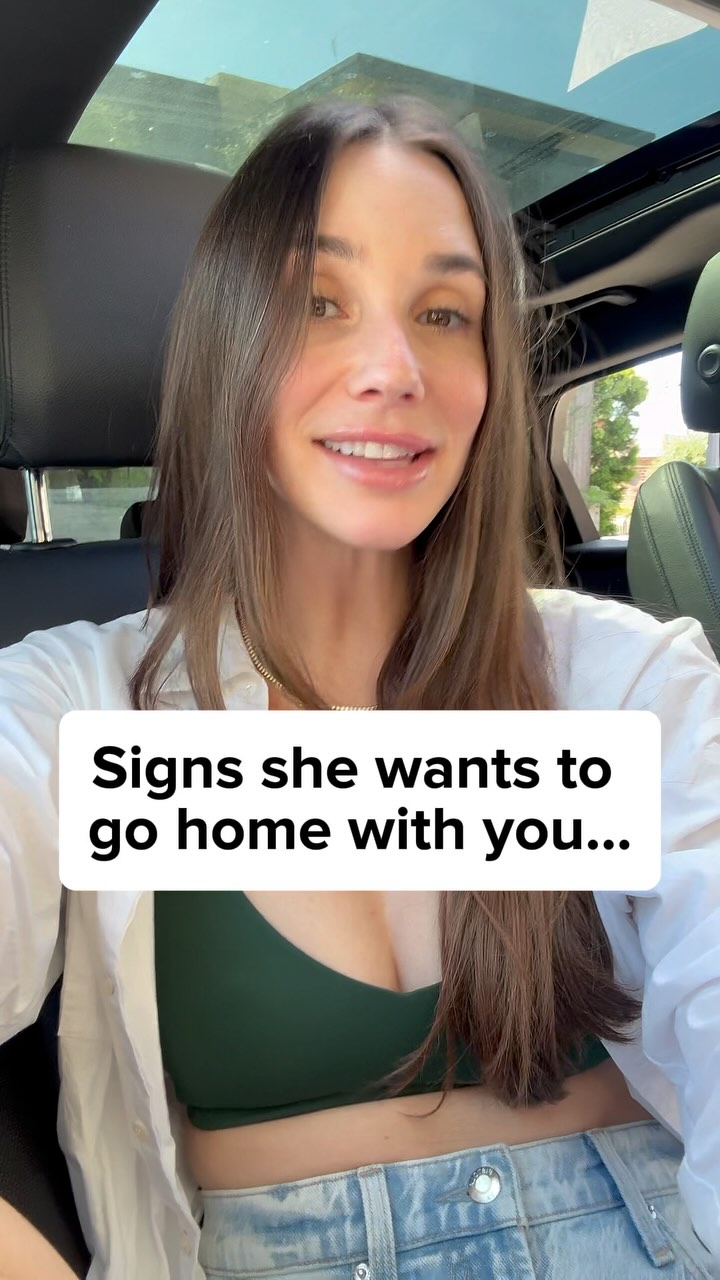 A woman in a car is smiling at the camera. She
looks to be in her vehicle, with a sign that reads,
"Signs she wants to go home with you..." in a
white box.