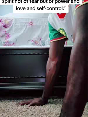 The image shows the lower portion of a person doing a push-up. The person is viewed at an angle, with only their arms, hands, and a portion of their torso visible, positioned near the ground with the hands placed on the floor for a push-up. The setting appears to be a room with a beige carpet. Behind the person, a bed is discernible, covered with a light-colored bed sheet. The lighting is moderate, with shadows indicating the direction of the light source. The scene conveys a sense of effort and physical activity.