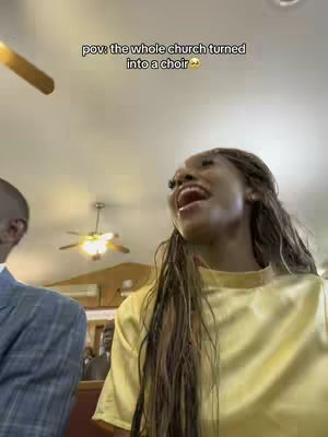 A woman with long, braided hair sings with her mouth open wide. She's wearing a yellow top, and a man in a suit is partially visible on the left. The background includes a ceiling, lights, and another figure, suggesting a church setting. Text overlay reads, 'pov: the whole church turned into a choir'.