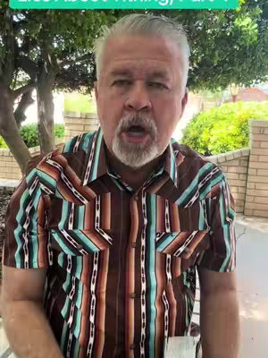 A man with gray hair and beard is speaking to the camera. He is wearing a patterned button-down shirt. The background consists of green foliage and a brick wall.
