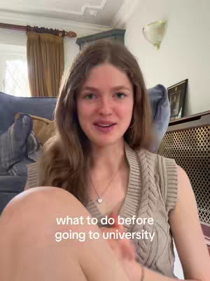 A young woman with long brown hair sits in front of a blue couch, looking at the camera and talking. Text overlaid on the image reads "what to do before going to university". The background includes window curtains and a wall sconce.