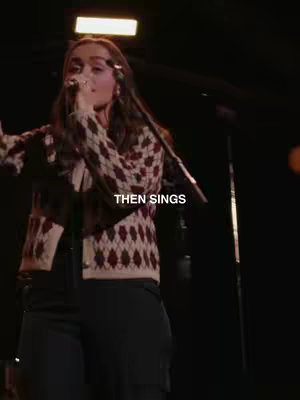 A woman singing into a microphone on a stage with dim lighting. She is wearing a patterned sweater and dark pants. The text overlay reads "THEN SINGS".