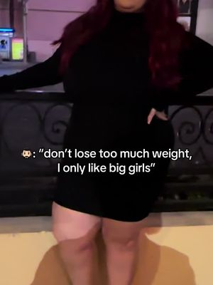 The image shows a woman in a black dress standing in front of a decorative metal railing. She has long auburn hair and is positioned from the mid-thigh up, showing the front of her torso. Text is overlaid at the bottom with a chat box, reading ,"don't lose too much weight, I only like big girls". The overall tone is casual, set in an outdoor location at night, with the suggestion of an active social setting.