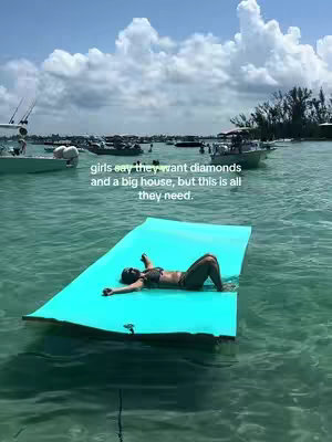 The image shows a woman lying on a bright blue floating mat in the ocean under a partly cloudy sky, surrounded by boats in the distance, capturing an idyllic, relaxed moment.