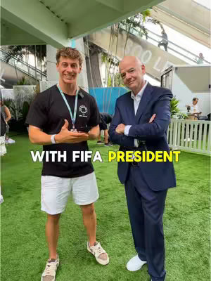 Two men stand on green grass under a metallic structure. The man on the left, wearing a black t-shirt and white shorts, is holding something while the older man on the right, in a suit, has his arms crossed. Text overlays say 'WITH FIFA PRESIDENT' in large yellow letters.