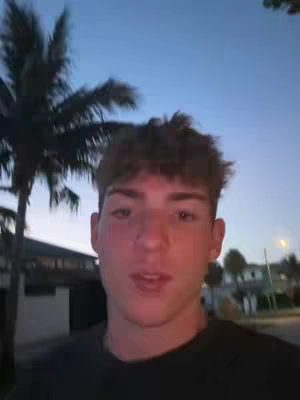 A young man stands outdoors, facing the camera. He has short, tousled brown hair and a neutral expression. The background includes a palm tree, a building, and a dusky sky, suggesting an early evening setting.