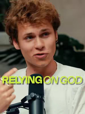 A young man with reddish-brown curly hair is centered in the frame, speaking with a microphone in front of him. He is wearing a white t-shirt and a small earring is visible on his left ear. The background is blurred. Overlaid on the image is text in yellow, reading 'RELYING ON GOD'.