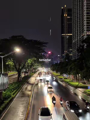 A nighttime cityscape with a wet road reflecting the lights of cars and streetlights. Tall buildings, a central road with traffic, and trees line the sides. The sky is dark, with a few streaks of light.