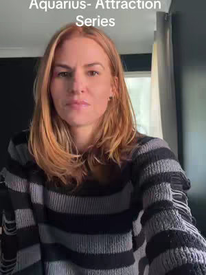 A woman is centered in the frame, facing the camera with a somber expression. She has fair skin, and her auburn hair is styled with a side part. She wears a black and gray striped sweater. The background appears to be an interior, with a dark wall and a partially visible curtain.