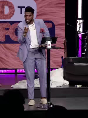 A man in a light purple suit stands on a stage, pointing towards the viewer. The background is a pink, textured wall with indistinct white letters. A table with a laptop and microphone is in front of him. 