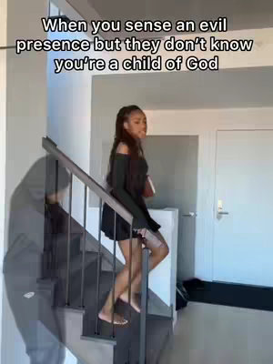 A woman walks down a flight of stairs inside a building, likely a home. The stairs are made of wood and have metal railings. The woman, dressed in a dark top and skirt, is the central focus and positioned on the stairs, suggesting movement. The image has a bright, well-lit appearance.