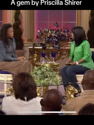 The image shows two women seated on chairs, engaged in conversation. The background features a decorated stage with floral arrangements and what appear to be audience members. The lighting suggests a studio setting.