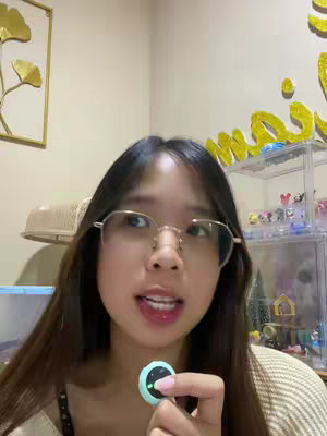 A young woman with long dark hair and glasses is centered in a room. She is wearing a light-colored, open-weave sweater and holding a small, round, teal-colored device in front of her. She has a slight smile, and her mouth is open as if she is speaking. Behind her is a cream-colored wall, with a golden leaf-shaped decoration. Above her shoulder is a decorative shelf on which small figurines are arranged. The lighting is warm, and the overall composition suggests a personal and intimate setting.