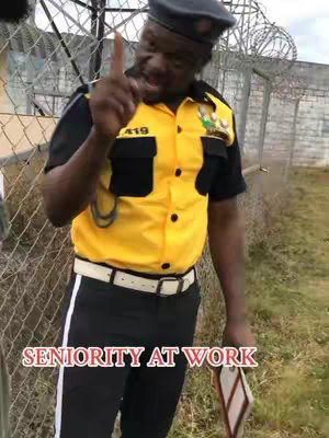 A man in a yellow and black uniform stands in front of a chain-link fence, holding something small in his right hand and pointing with his index finger upwards. Text overlayed at the bottom reads "SENIORITY AT WORK".