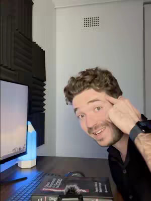 A person is inside a room with light gray walls and the room is lit by artificial lighting. The creator is smiling, pointing to their head near the right side, with a monitor on the left. A table is in front of the creator with a keyboard, book, and pen light. The background is a wall with acoustic panels on the left corner.