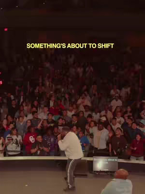 The image shows a large gathering of people in an auditorium, facing a stage. A man in a light-colored shirt is speaking into a microphone on the stage. The text overlay says 'Something's About to Shift'.