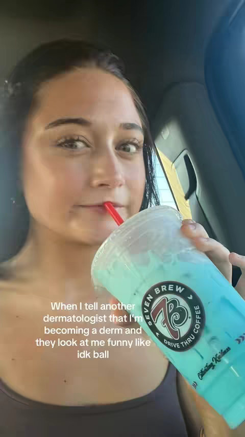 A young woman is seated in a car, seen from the chest up, facing towards the camera. She has dark hair that is pulled back, and she is wearing a tank top. She has a drink with a blue liquid and a red straw. Overlay text reads: When I tell another dermatologist that I'm becoming a derm and they look at me funny like idk ball. The background seems to be the inside of a car with the sun shining in, creating warm tones and highlighting the woman's face.