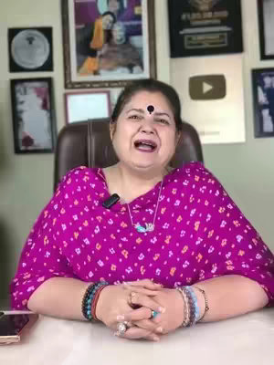 A woman with a bindi sits at a desk with her hands clasped. She wears a magenta top and multiple bracelets. Behind her are framed pictures, a YouTube play button, and a wall decoration.