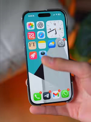 An iPhone held in a person's hand displays the home screen with multiple apps and a custom turquoise background. The camera is positioned to capture this content, with a hand holding the phone from the right. The scene is illuminated by natural light. The apps include a variety of social and utility icons. The phone is in a black case.