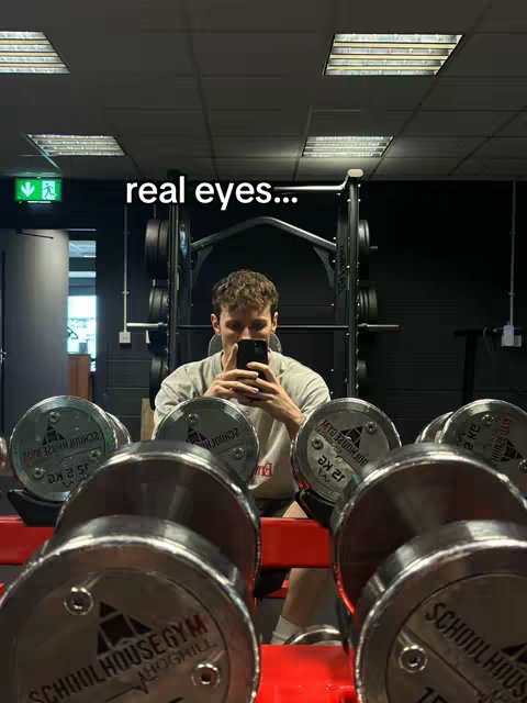 A man is taking a selfie in a gym, reflected in a large mirror. He is holding his phone up to take the picture. The background partially reveals gym equipment, including dumbbells and racks. Above the man, white text reads "real eyes..."