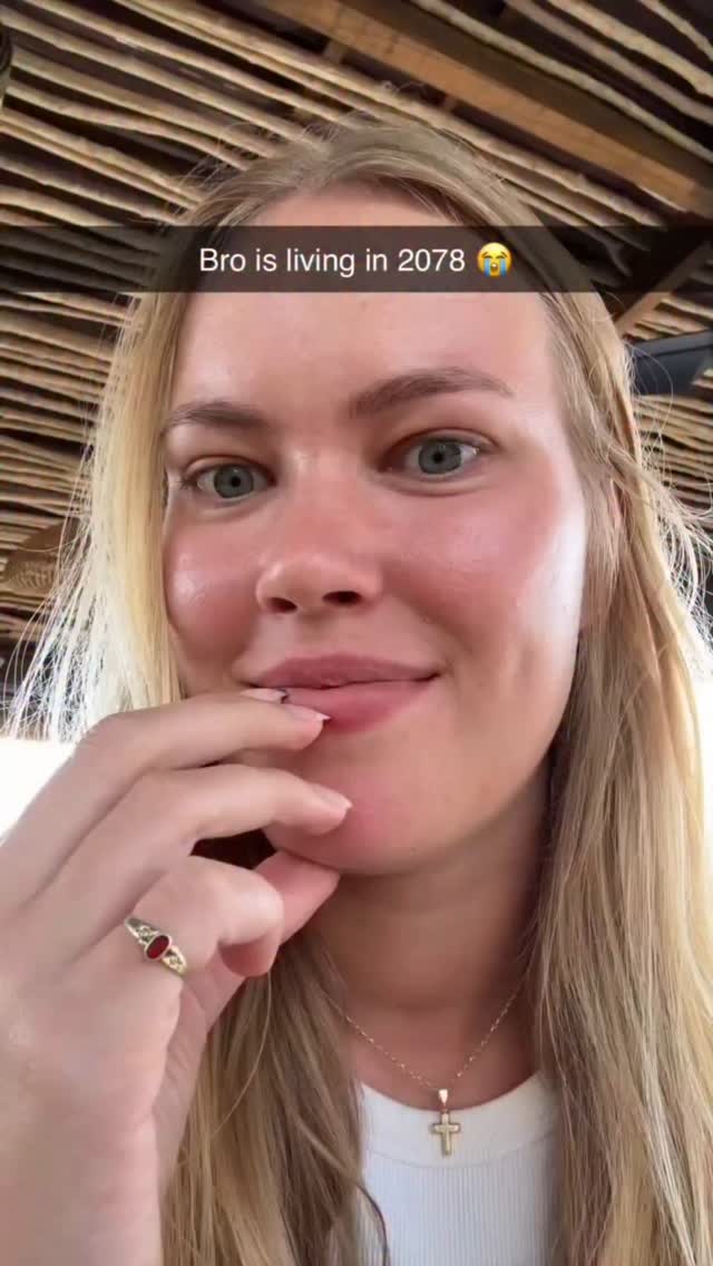 A close-up shot of a young woman with blonde hair, fair skin, and light-colored eyes. She is looking at the camera with a slight smile and has her finger close to her lips. The background is blurred and suggests an outdoor setting. A text overlay reads 'Bro is living in 2078'.