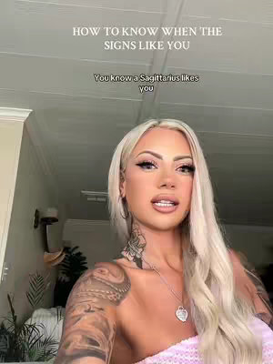 A woman with long, blonde hair is shown with her upper body, a large chest tattoos and a heart-shaped necklace . Overlay text at the top and in the middle informs viewers about astrological signs.