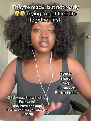 A woman with curly dark hair is centered in a selfie-style image, wearing a black tank top and white earbuds. Text overlays are present at the top and bottom, and text in a box to the right identifies astrological signs. The background is softly blurred, hinting at a domestic setting.
