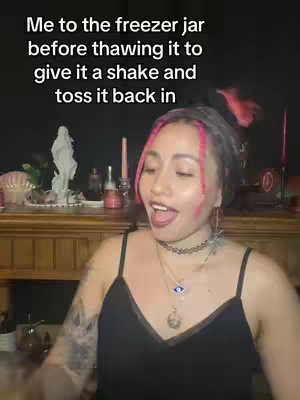 A woman with pink-streaked hair is the central focus of the image, mid-speech and framed above a wooden surface. A dark background sets the mood, and text overlays. Adjacent to the woman, floral elements and candles are arranged providing a witchy or spiritual ambience. 