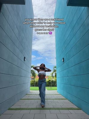 A woman stands facing the camera in a narrow passage between two tall blue walls, hands near her face. The background is a lush green area and a partly cloudy sky.