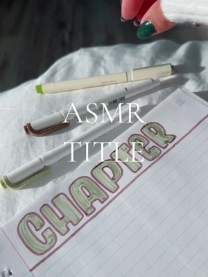 The image features a close-up shot of stationery on a white surface. Two white pens and a brown-tipped pen are laid on top of a notebook page, over which the words "ASMR TITLE CHAPTER" are handwritten in green pen. The scene is lit in natural light and the hand of a person with green nail polish holds and points at the design.