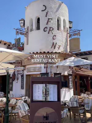 The image shows a restaurant facade on a sunny day. The restaurant has a white, rounded tower with the word "DIFERENT" printed in large letters. Below the tower, there's outdoor seating with tables and chairs under umbrellas. A sign is visible with the restaurant name and a menu board. The sky is blue and clear.