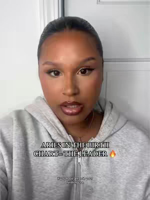 A medium shot of a woman with styled dark hair pulled back, wearing grey hoodie and makeup, against a white background. Text overlayed reads, 'ARIES IN THE BIRTH CHART- THE LEADER'. Additional text at bottom.