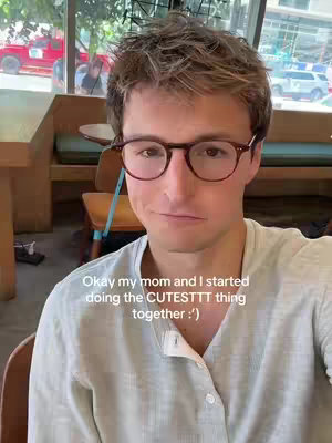 A young man with short brown hair and glasses is in the frame, the man is looking towards the camera and has a slightly raised eyebrow, he is wearing a light colored collared shirt, and behind the man a cafe setting with tables and chairs is visible.