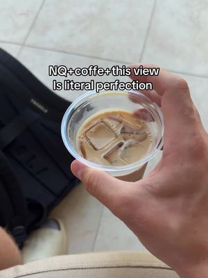 A selfie-style shot with a hand holding a clear plastic cup of iced coffee. The drink has ice cubes and a light brown coffee color. The background is blurred, with a partial view of a black backpack and a light-colored floor. The focus is on the hand and the drink, with the text overlay on top.