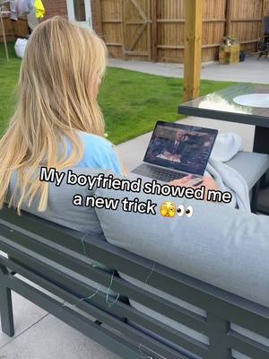 A woman with long blonde hair is sitting on an outdoor bench, looking towards a laptop on her lap. The screen displays an image of a man in a suit. Grass, a fence, and the corner of a building make up the background.