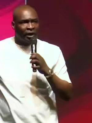 A medium shot shows a bald man, wearing a white shirt, holding a microphone and speaking. The background is a gradient of pink and red hues.