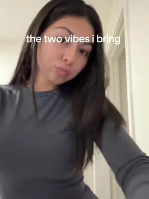 A woman is shown in a close-up selfie-style shot. She is looking slightly upward. She is wearing a dark gray long-sleeve shirt. The background is a neutral-colored wall with a door frame visible at the right.