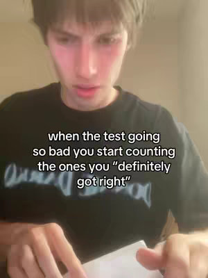 A medium shot of a young man looking down at a paper, likely a test or exam. He has short, dark hair and is wearing a dark t-shirt. Text overlayed on the image gives context to the situation, implying a challenging or stressful academic experience.