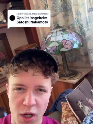 A young man with short, curly hair and a dark cap is looking at the camera. A speech bubble indicates the comment 'Opa ist insgeheim Satoshi Nakamoto'. The background includes a decorative lamp, a bookshelf and some other blurred objects adding to the context.