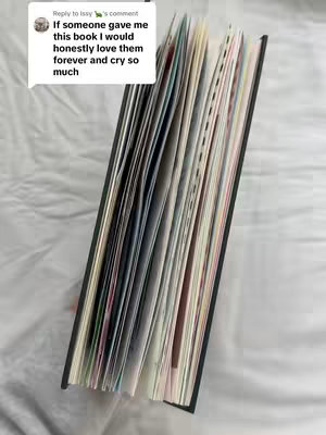 An open book is seen at a slight angle, showcasing numerous colorful pages. The book is resting on a white, textured surface, possibly a bed sheet or blanket. In the upper left corner, there's a comment overlaid on the image, and it presents text related to the book.