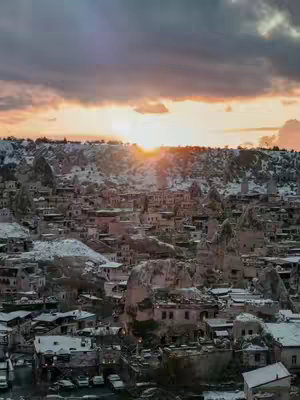 A scenic landscape shot featuring a town nestled within a valley, with unique rock formations and buildings. The sky shows a sunset or sunrise with warm, orange hues, partially obscured by clouds. The buildings have a uniform appearance. The overall effect suggests a chilly, possibly winter, atmosphere.