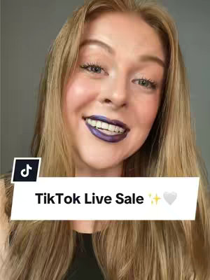 A medium shot features a fair-skinned woman with long, light-blonde hair, smiling at the camera. She has full lips painted with a deep purple lip color and is wearing a black top. A text overlay near the bottom reads 'TikTok Live Sale' with a sparkle and a heart emoji. The background is a neutral gray.
